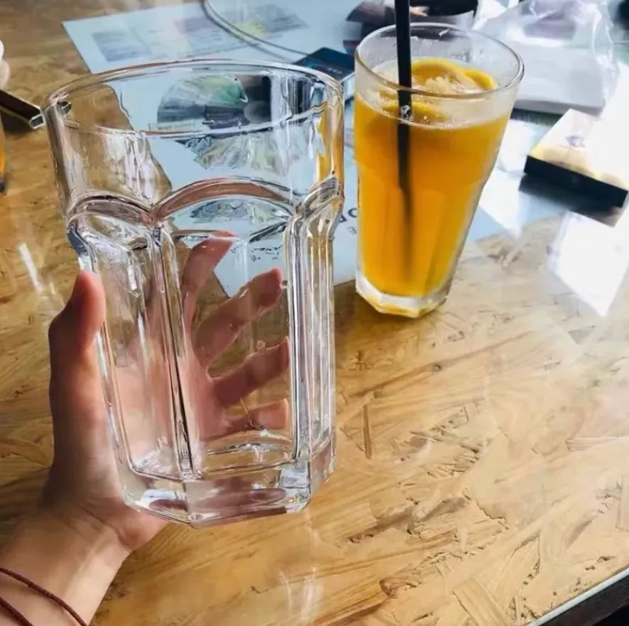 Verre à cocktail Mojito XL tenu en main, vide et transparent, à côté d'un jus d'orange glacé avec paille