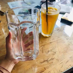 Verre à cocktail Mojito XL tenu en main, vide et transparent, à côté d'un jus d'orange glacé avec paille