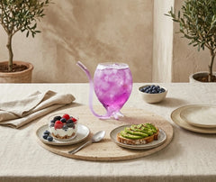 Verre à cocktail paille posé sur une table de brunch méditerranéen avec yaourt aux fruits rouges, tartine à l'avocat et oliviers en pot