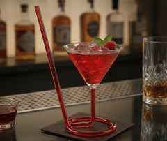 Verre à cocktail avec paille intégrée rose, rempli d'un cocktail rose garni d'un demi fruit de la passion, sur fond rouge bordeaux avec plateau cuivré