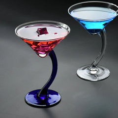 Deux verres à cocktail pied courbé côte à côte, l'un avec un cocktail rouge sur pied bleu cobalt, l'autre avec un cocktail bleu sur pied transparent, sur fond sombre.