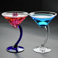 Deux verres à cocktail pied courbé côte à côte, un cocktail rouge sur pied bleu cobalt et un cocktail bleu sur pied transparent, vue d'ensemble sur fond gris clair.