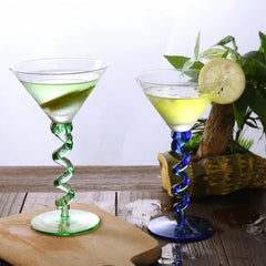 Les verres à pied torsadé vert et bleu remplis de cocktails, posés sur une planche en bois avec une plante verte en arrière-plan.