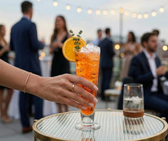 Une main tient un verre à cocktail Pilsner rempli d'un spritz orange garni d'une rondelle d'orange et de thym, posé sur une table ronde miroir lors d'une soirée rooftop au coucher du soleil.