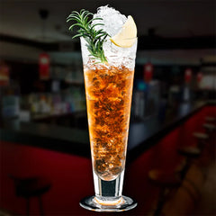 Verre à cocktail Pilsner rempli d'un cocktail ambré glacé, garni d'un brin de romarin et d'un quartier de citron vert, sur fond de bar flouté.