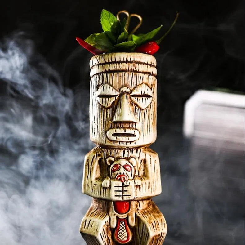 Gros plan du visage sculpté du verre tiki, surmonté de menthe fraîche et de piments rouges, dans une ambiance fumée.