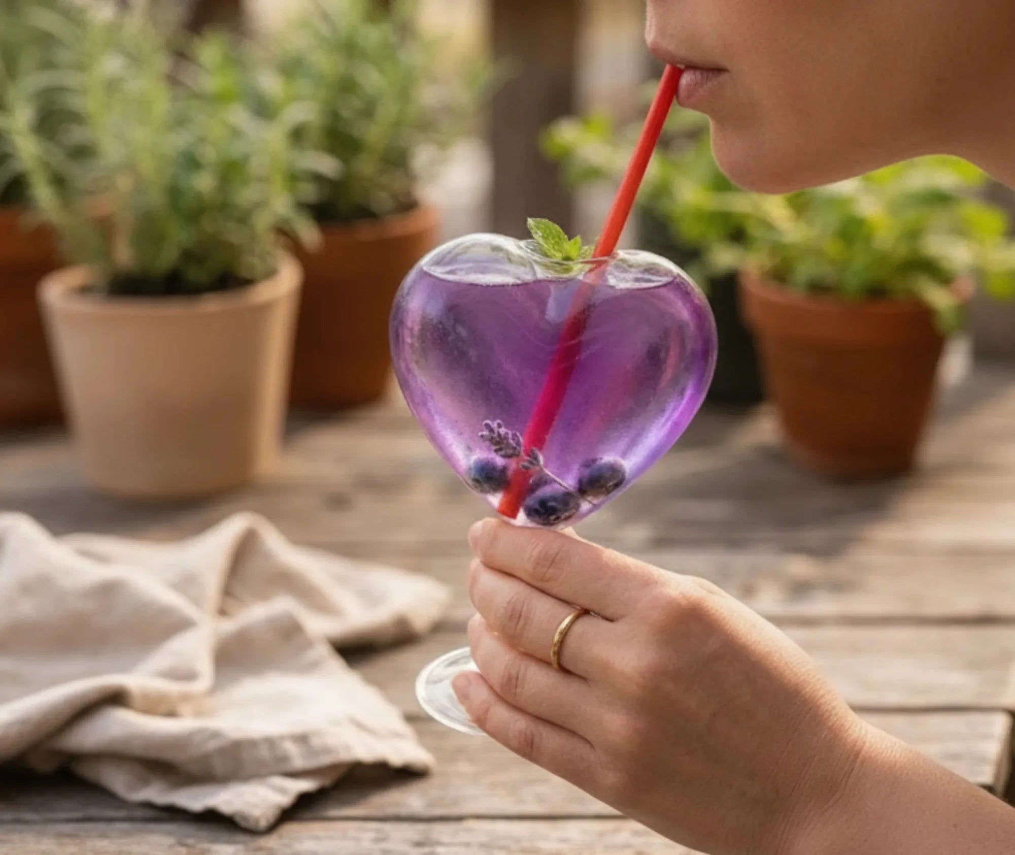 Une femme déguste avec une paille rouge un cocktail violet aux myrtilles servi dans un verre en forme de cœur, en terrasse