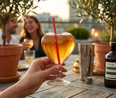 Une main tient un verre à cocktail en forme de cœur rempli d'un cocktail doré, sur une terrasse au coucher du soleil