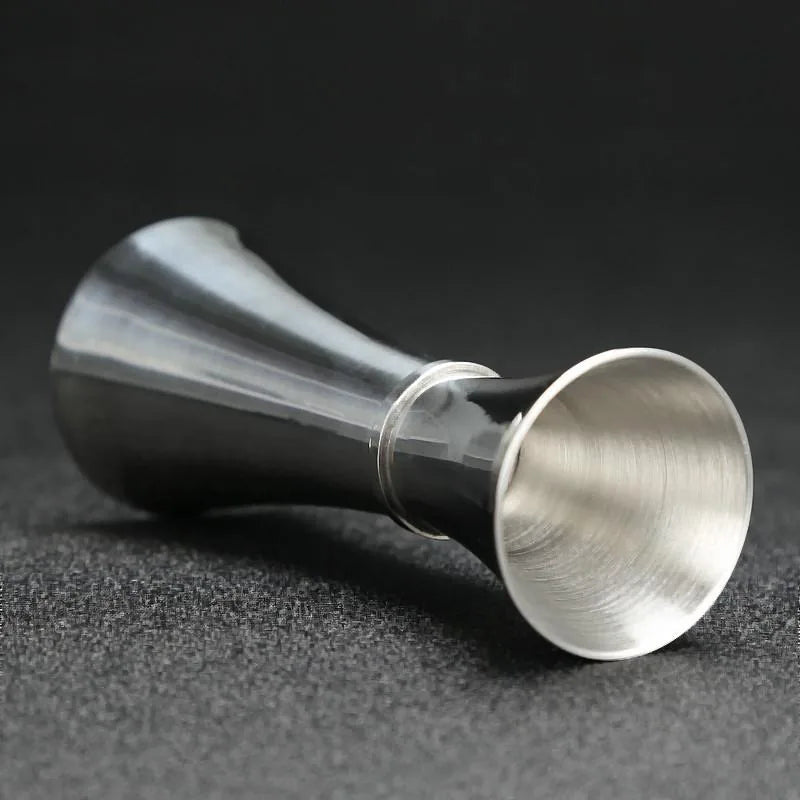 Verre doseur à cocktail en inox couché sur le côté, vue rapprochée sur fond gris foncé.