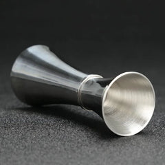 Verre doseur à cocktail en inox couché sur le côté, vue rapprochée sur fond gris foncé.