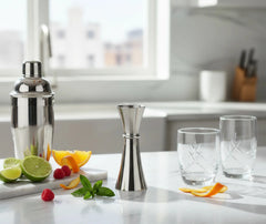Verre doseur à cocktail posé sur un plan de travail en marbre, entouré d'un shaker, de citrons verts, d'oranges, de framboises et de menthe fraîche.