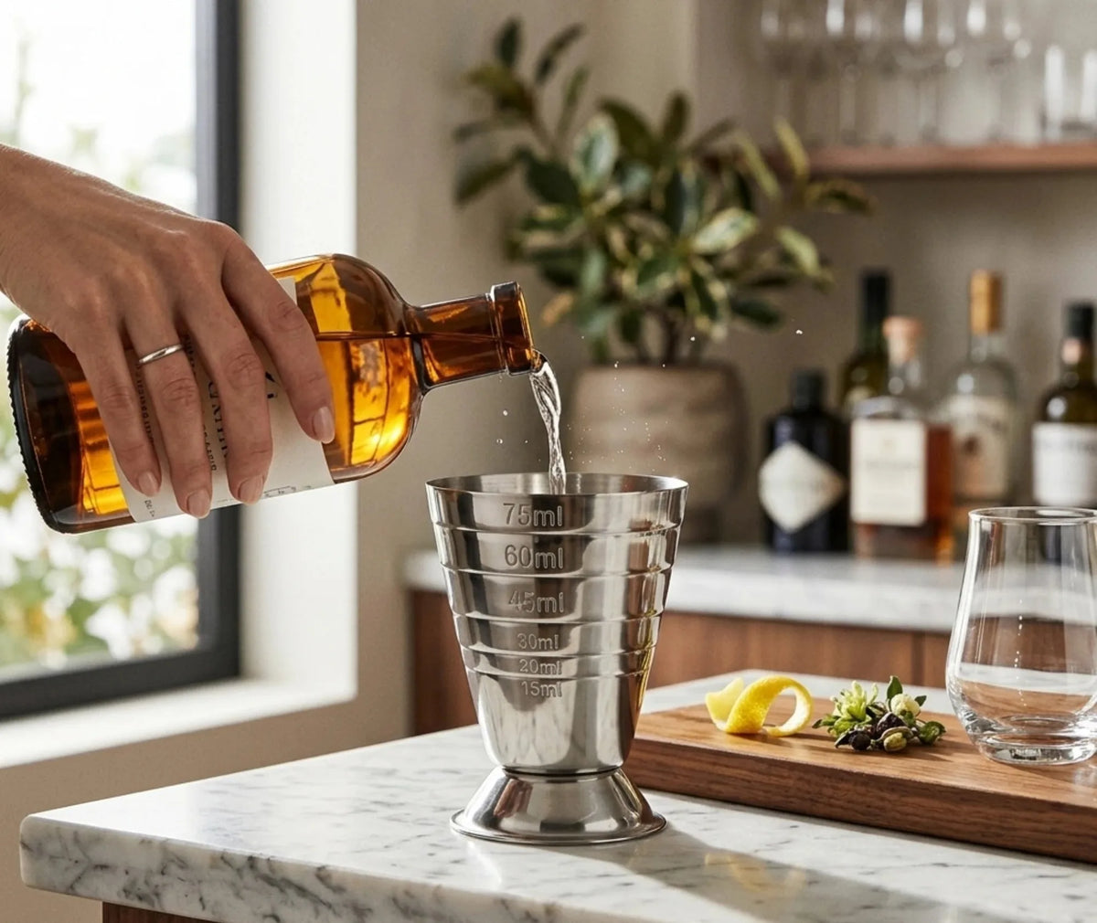 Une main verse une liqueur ambrée dans un verre doseur à cocktail posé sur un plan de travail en marbre, avec écorce de citron et baies à proximité.