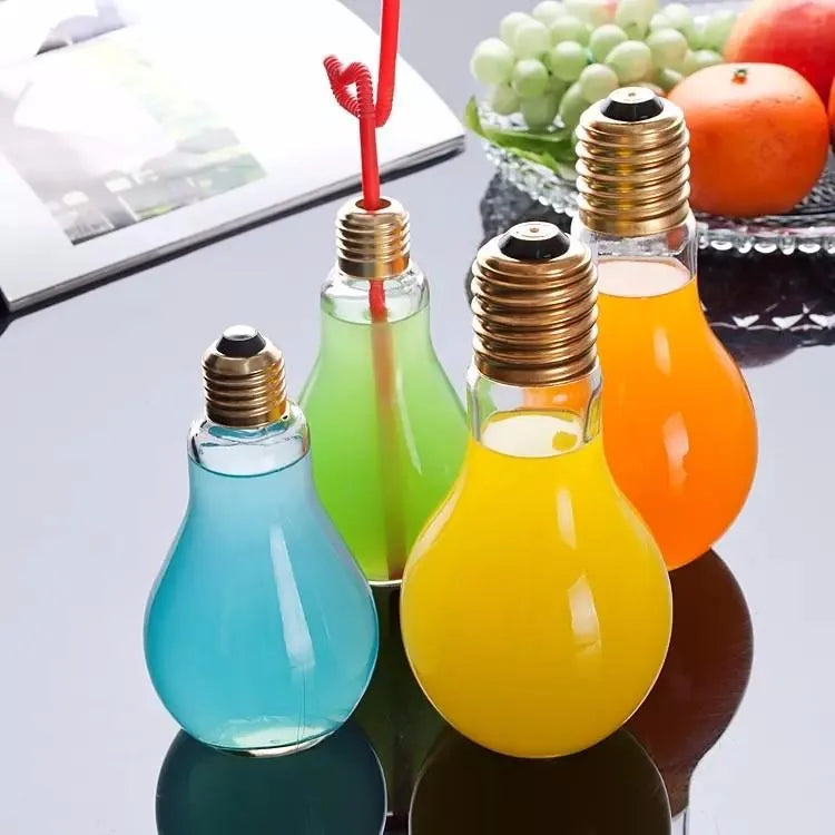 Quatre verres à cocktail en forme d'ampoule remplis de boissons bleue, verte, jaune et orange, avec une paille rouge, posés sur une table à côté d'un magazine et d'une coupe de fruits frais.