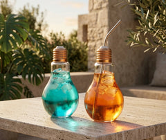 Deux verres à cocktail en forme d'ampoule remplis de boissons turquoise et ambrée, garnis de glaçons et d'une paille métallique, posés sur une table en pierre en extérieur au coucher du soleil.