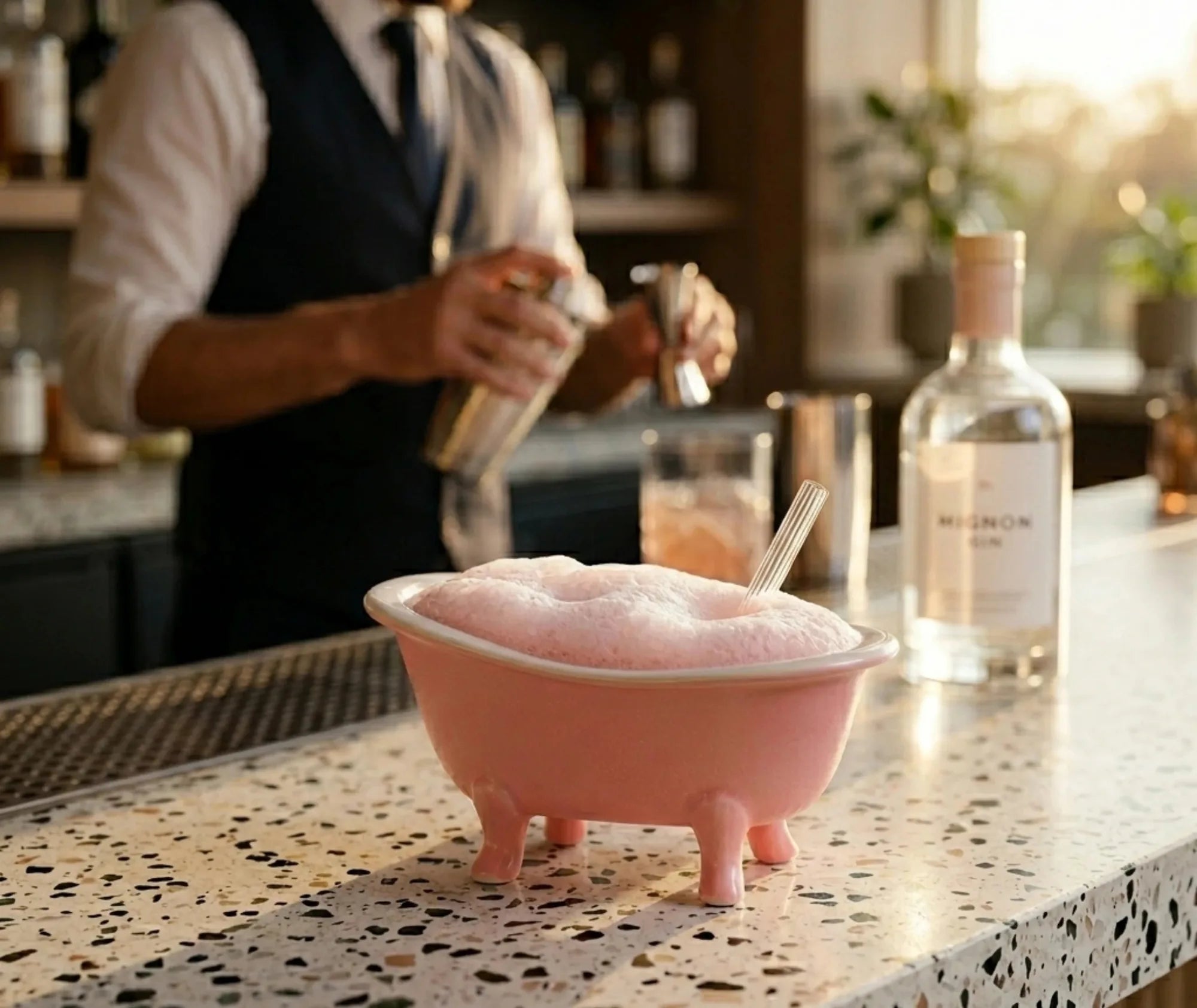 Verre cocktail en forme de baignoire rose rempli de mousse, posé sur un comptoir de bar avec un barman préparant un cocktail en arrière-plan