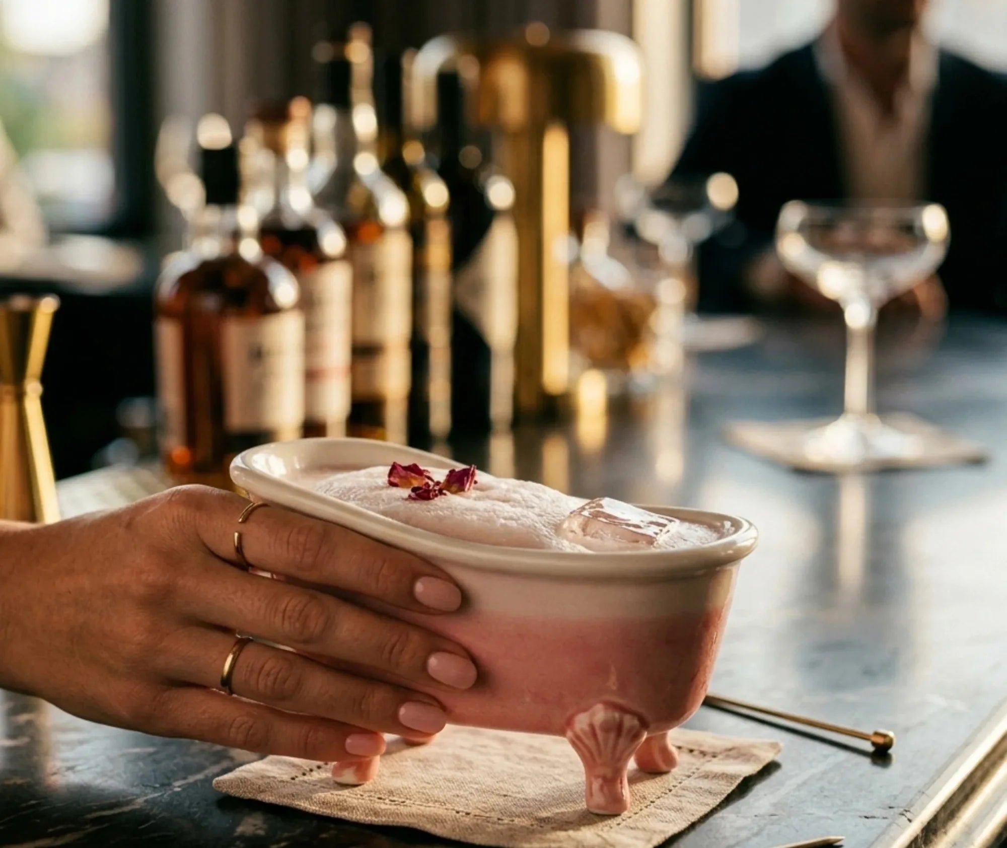 Main tenant un verre en forme de baignoire rempli de mousse, d'un glaçon et de pétales de rose, dans une ambiance de bar chic