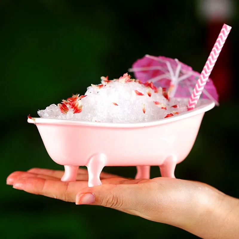 Verre en forme de baignoire rose tenu dans une main, rempli de glace pilée et de pétales, avec un parasol de cocktail rose