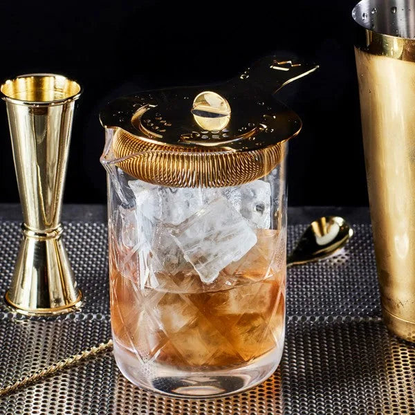 Verre mélangeur avec passoire à julep dorée posée dessus, cocktail ambré et glaçons, accessoires de bar en arrière-plan