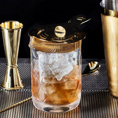 Verre mélangeur avec passoire à julep dorée posée dessus, cocktail ambré et glaçons, accessoires de bar en arrière-plan