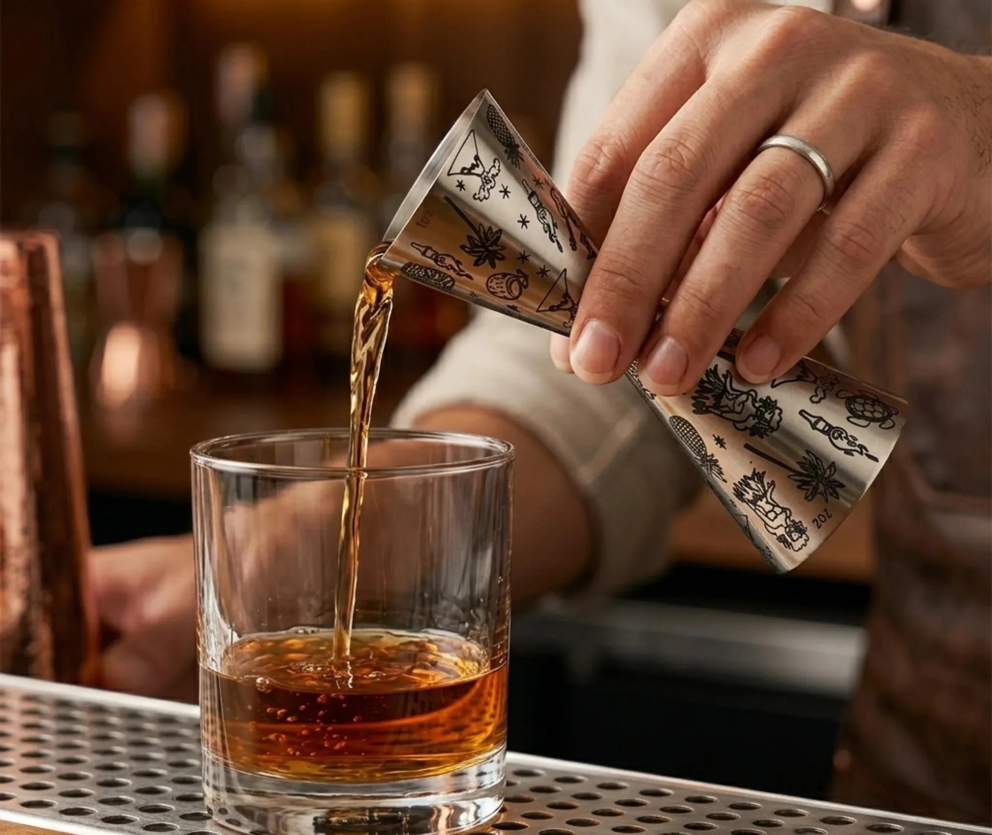 Un barman verse un cocktail ambré dans un verre à fond épais à l'aide du verre mesureur à motifs tropicaux, au comptoir d'un bar.