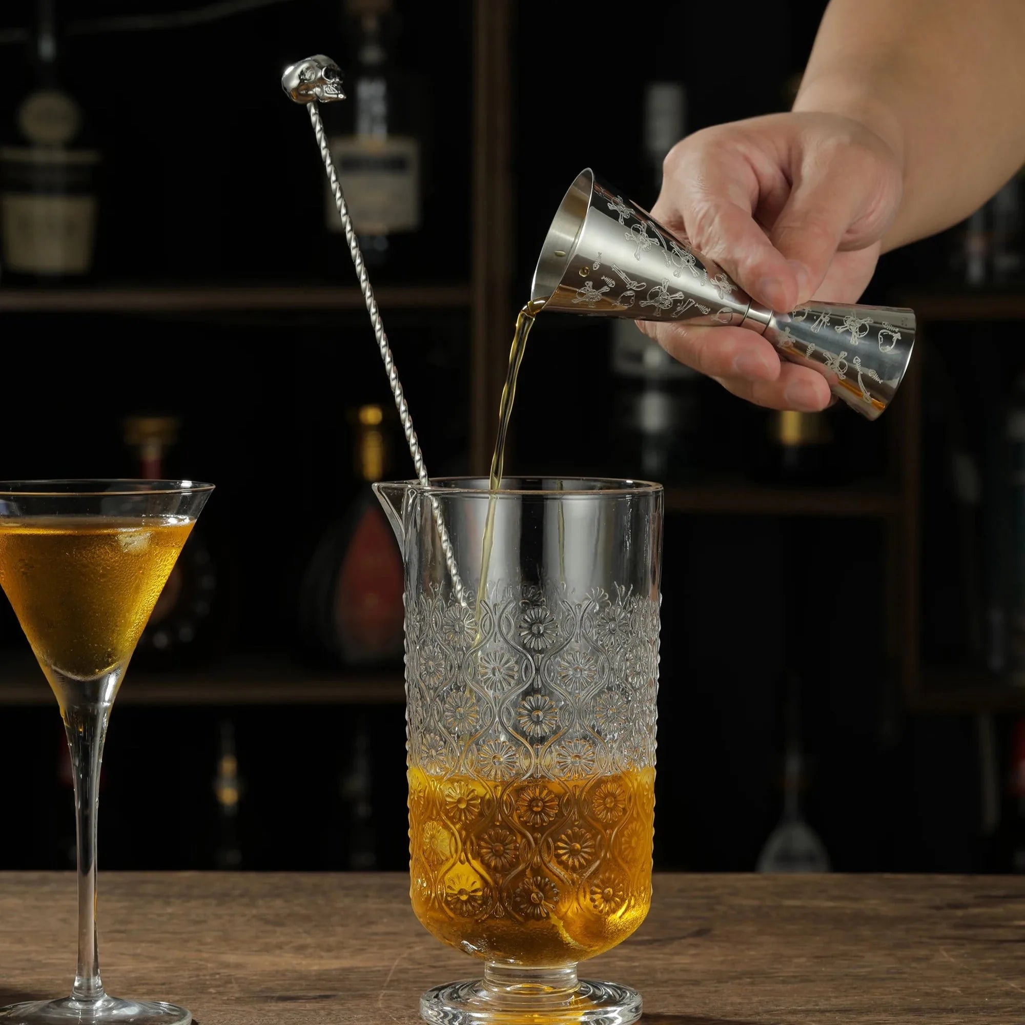 Une main verse un cocktail avec le verre mesureur à motif tête de mort dans un verre à mélange, près d'une cuillère à cocktail surmontée d'un crâne.