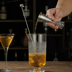 Une main verse un cocktail avec le verre mesureur à motif tête de mort dans un verre à mélange, près d'une cuillère à cocktail surmontée d'un crâne.