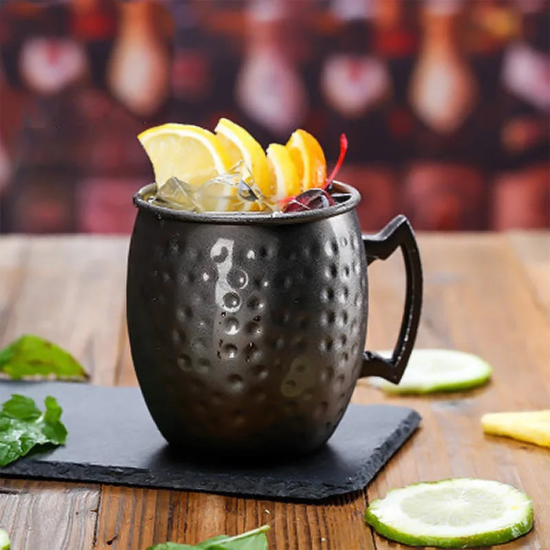 Verre à cocktail Moscow Mule noir garni de tranches de citron et d'orange et de glaçons, posé sur une ardoise dans une ambiance de bar.