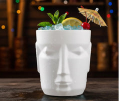 Gros plan sur le verre tiki blanc garni de menthe, citron, cerise et ombrelle en papier, posé devant des lumières de bar floutées