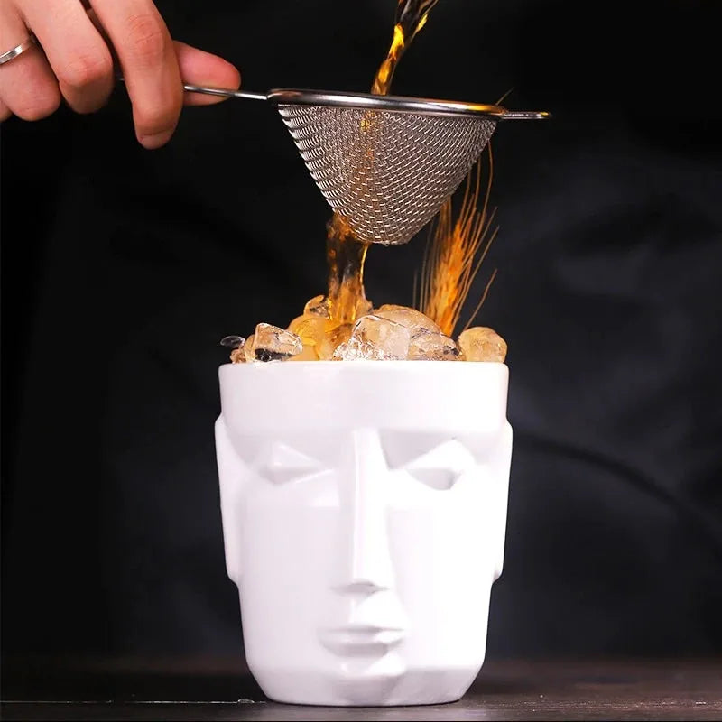 Un cocktail ambré est versé à travers une passoire fine directement dans le verre tiki blanc rempli de glaçons, sur fond sombre
