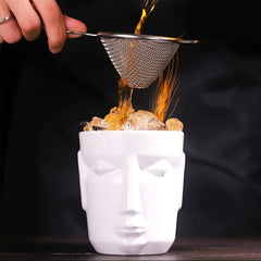 Un cocktail ambré est versé à travers une passoire fine directement dans le verre tiki blanc rempli de glaçons, sur fond sombre