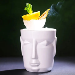 Le verre tiki blanc garni de citron et de fruit séché dégage une légère fumée, mis en scène sur fond vert foncé