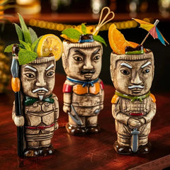 Trois verres à cocktail Tiki Suavecita aux visages de guerriers sculptés, garnis de menthe, d'agrumes et d'un parasol en papier, posés sur une table en bois sombre.