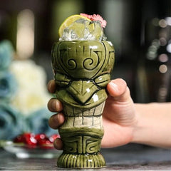 Gros plan du verre tiki vert rempli de glaçons, garni d'un quartier de citron et d'une fleur rose, tenu à la main.