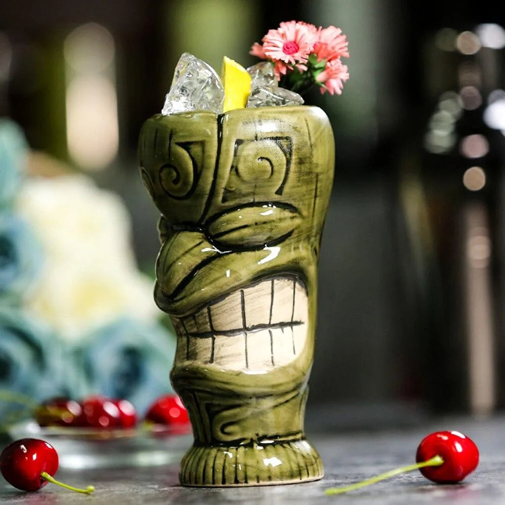 Le verre tiki vert posé sur une table en pierre, entouré de cerises fraîches, avec des glaçons et une fleur rose à l'intérieur.