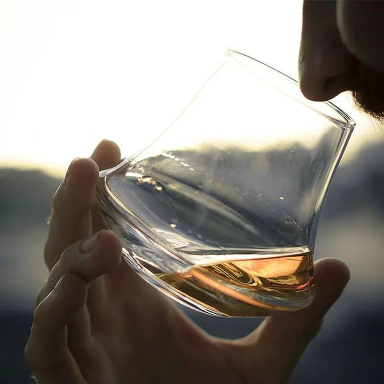 Gros plan d'une personne dégustant du whisky dans le verre tulipe moderne, en extérieur à la lumière du coucher de soleil.