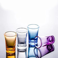 Quatre verres à shot de teintes différentes (orange, transparent, bleu, violet) posés sur une surface réfléchissante.
