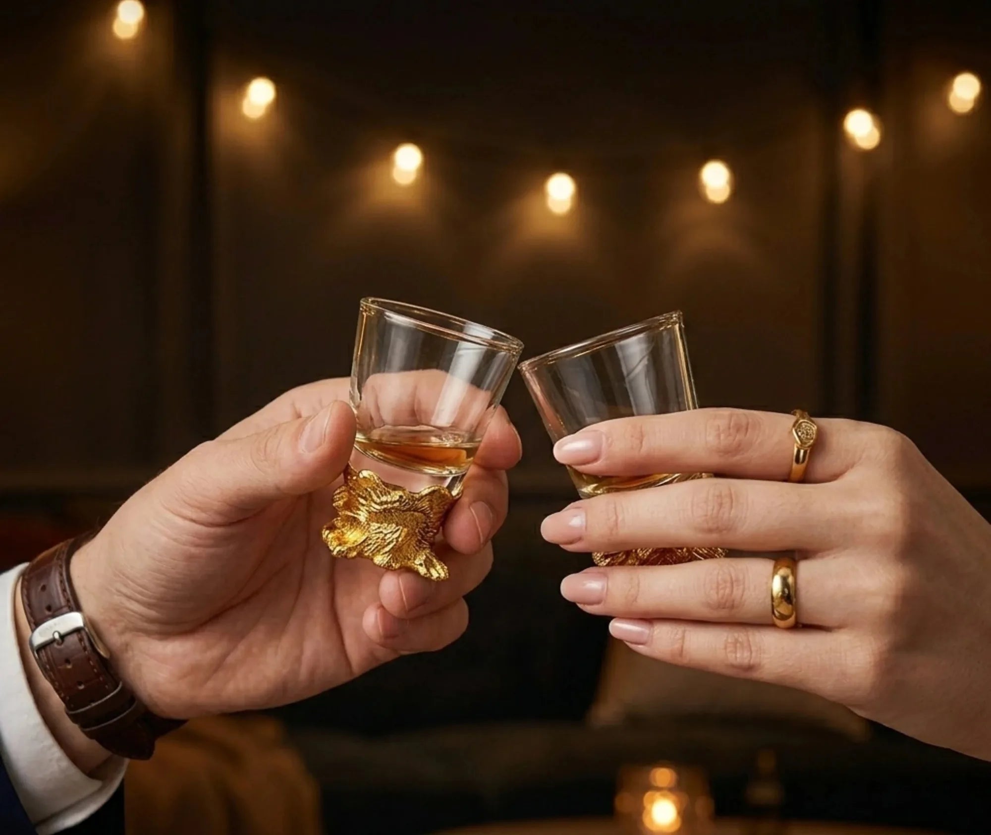 Un couple trinque avec deux verres à shot ornés de têtes d'animaux dorées, ambiance bar chaleureuse avec lumières en arrière-plan.