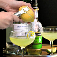 Zesteur à agrumes en train de zester un citron au-dessus d'un cocktail en coupe, avec bouteilles de spiritueux en arrière-plan.